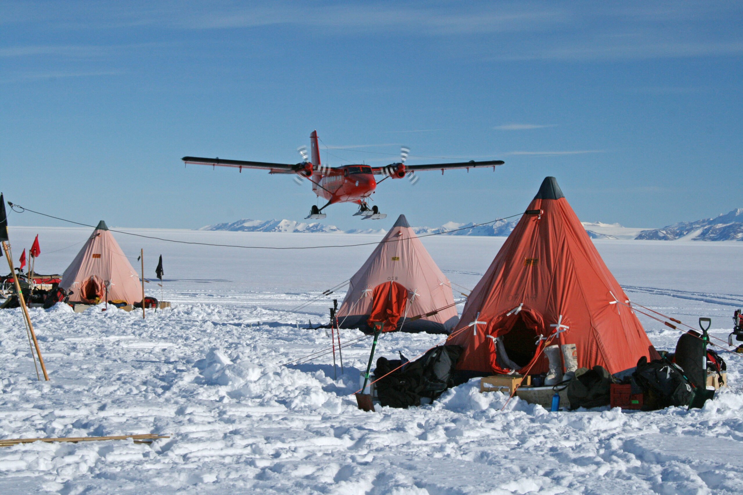 Three orange pyramid tents in a snowy landscape, with a red plane landing in the background.