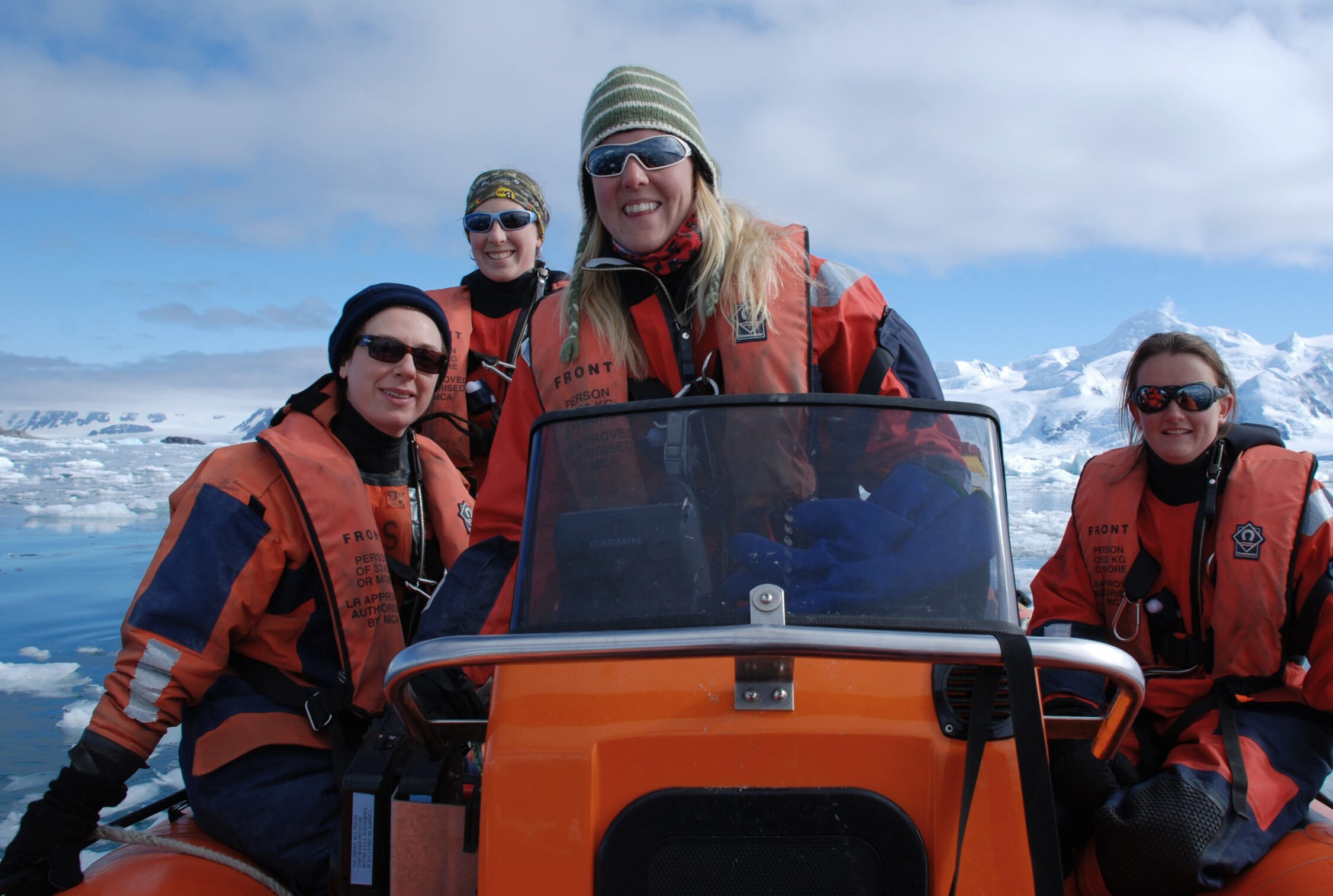 Four women wearing sunglasses and hats are sat on a boat, smiling. They are wearing orange boat suits and life jackets. There are snow covered mountains in the background.