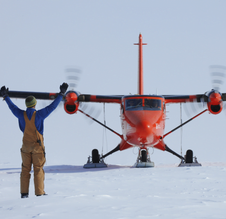 A man in a blue jumper, cream dungarees, a hat and gloves stands with his hands in the air, in front of a red plane landing on snow.