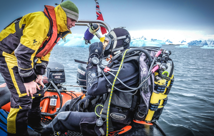 Two people on a boat. The man on the left is wearing a boat suit and life jacket and is checking the diving equipment of the person on the left who is wearing a drysuit and scuba diving gear, There are icebergs in the background.