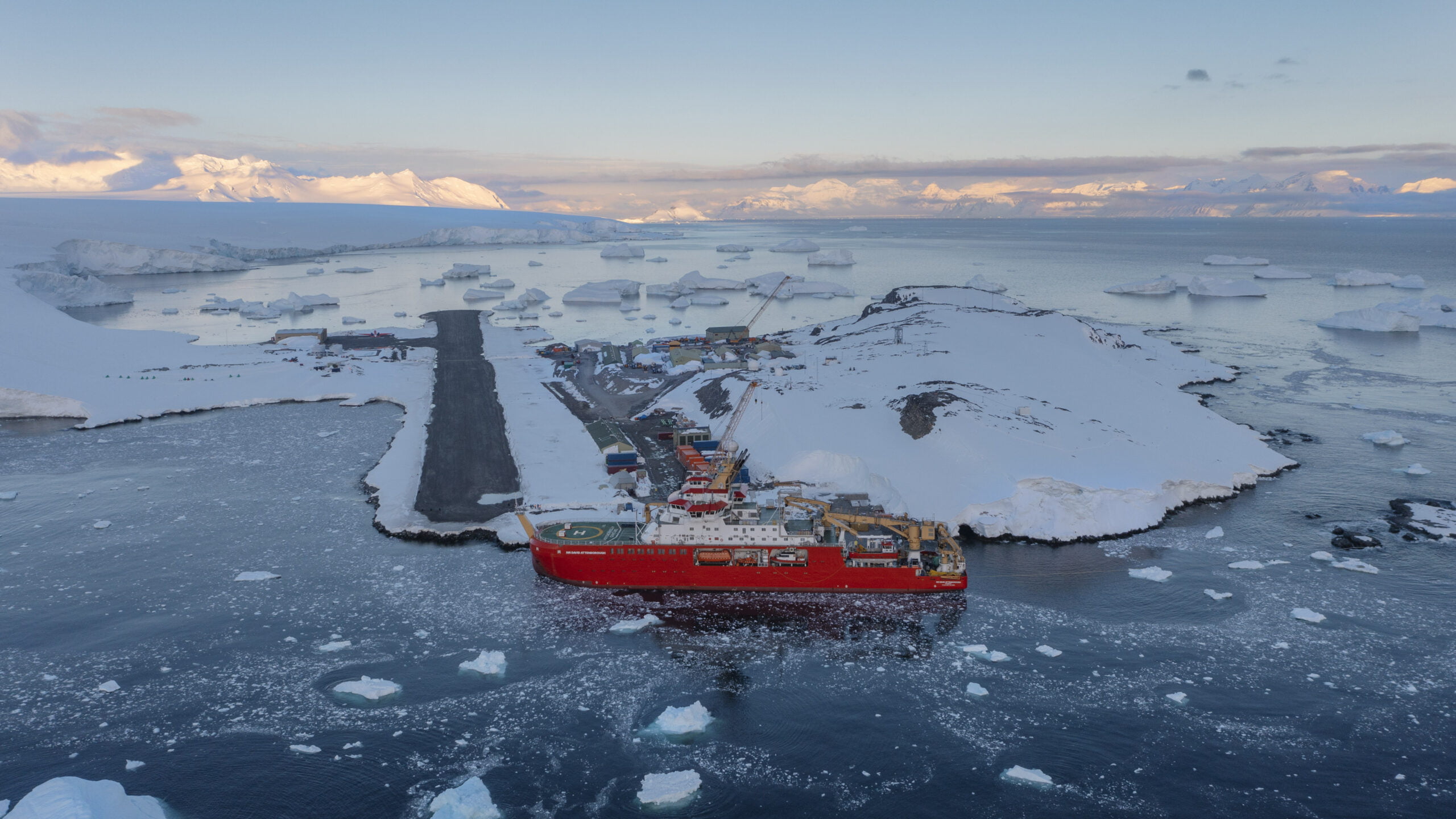 An aerial photo of a large red ship berthed up next to a snow-covered peninsula. There are bits of sea ice and icebergs in the water, and snow-covered mountains in the background.