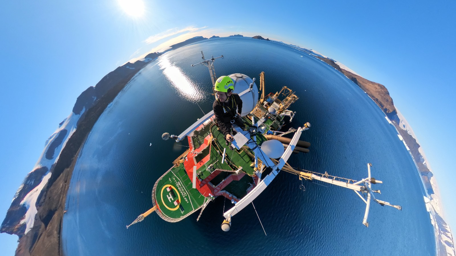 A fish-eye style photo of a man in a hard hat on a ship, surrounded by water 