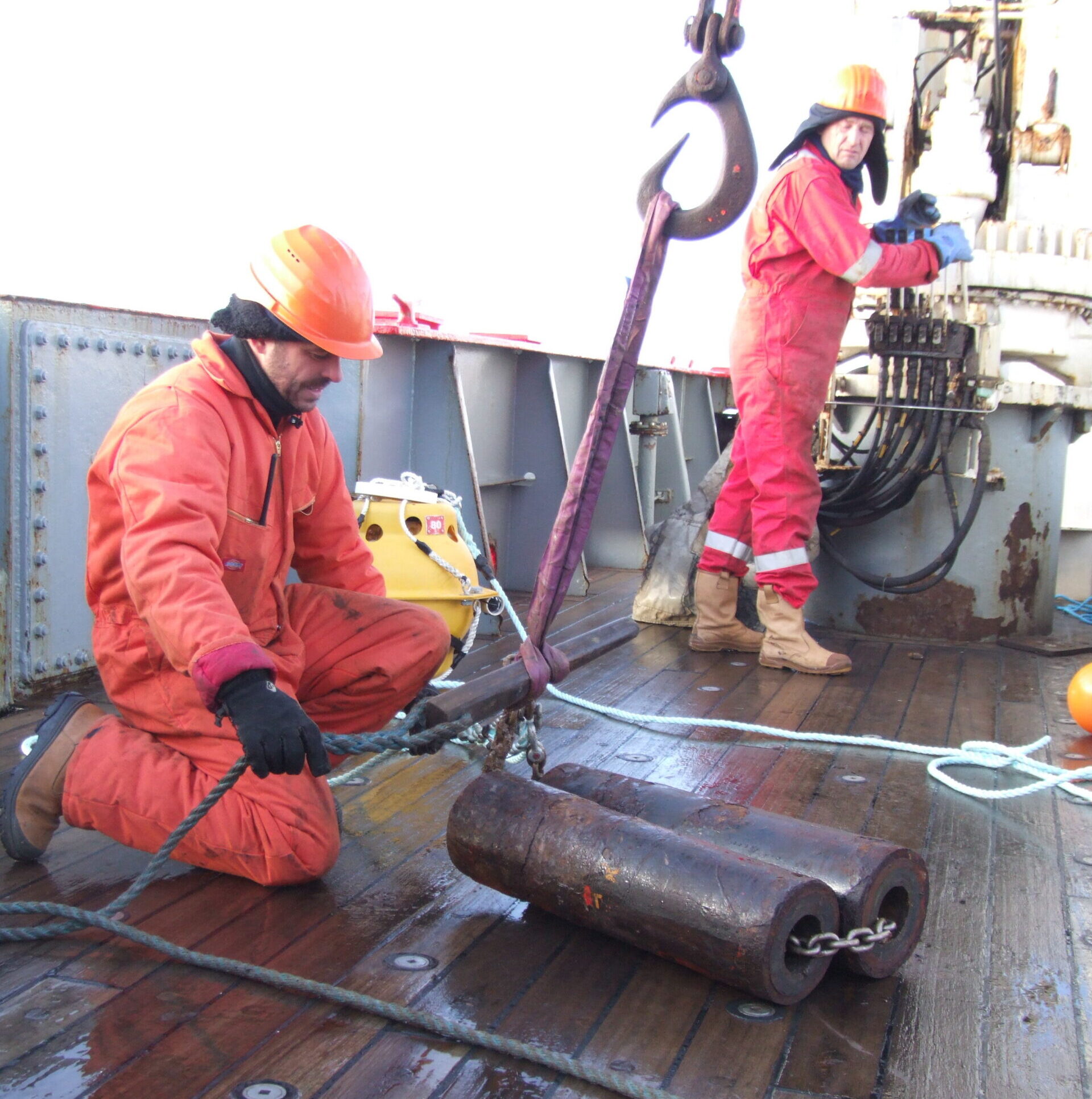 A man in an orange boiler suit and hard hat kneeling down on the deck of a ship, holding a rope