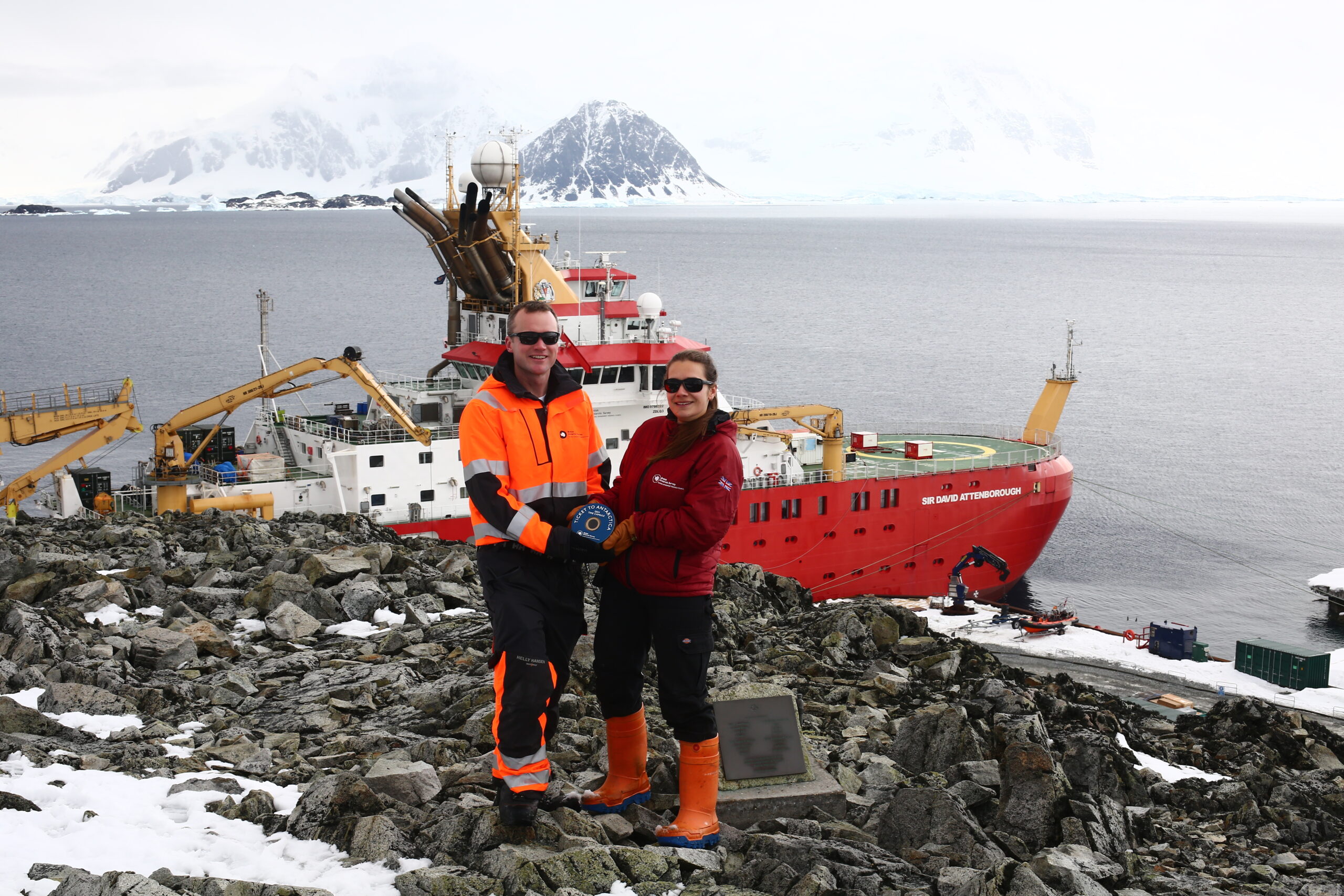 Two p[eople standing on a hill, holding a time capsule with a ship in the background