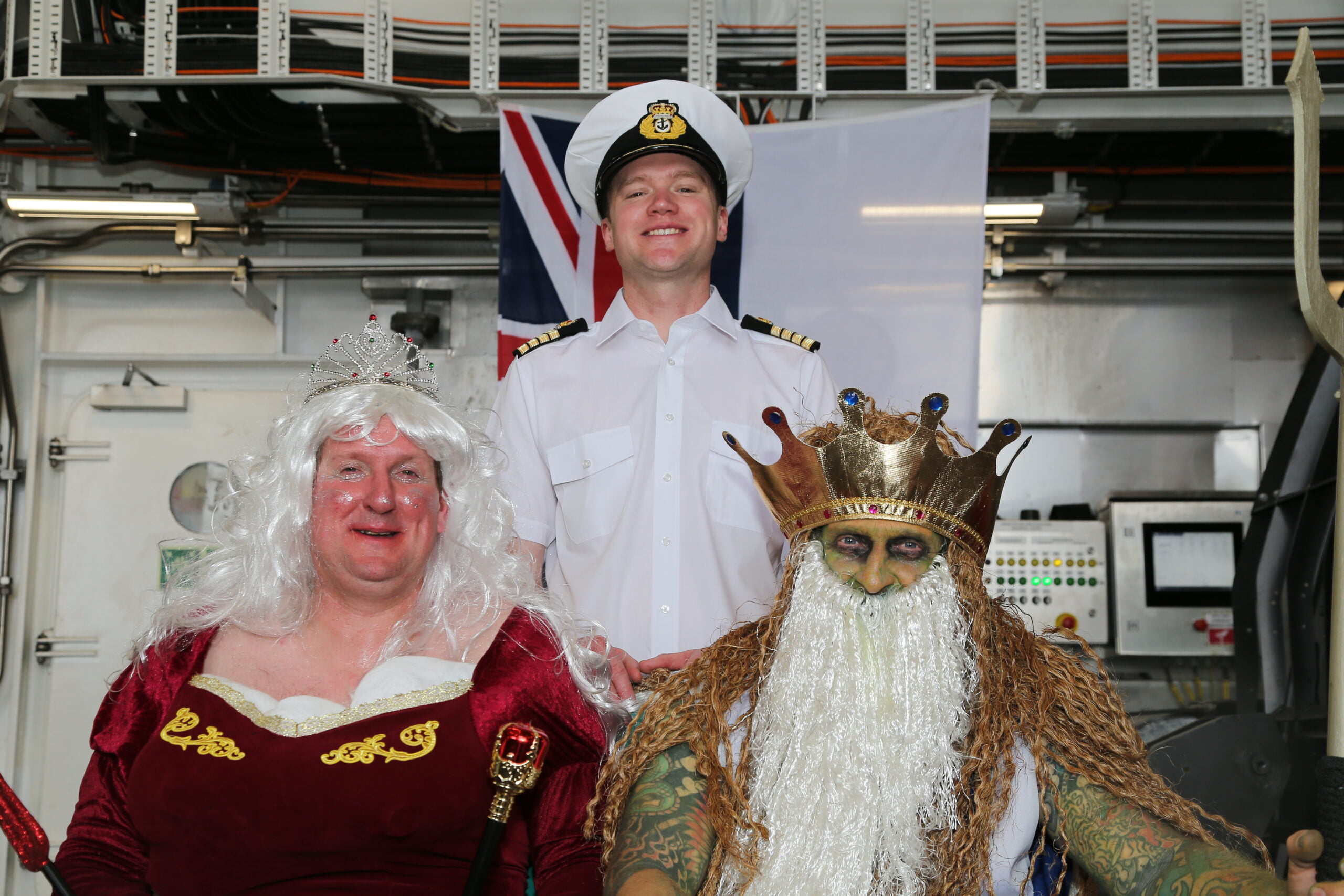 The Captain of the ship stands behind two mean in fancy dress. One is dressed as King Neptune, with a crown and beard. Another is dressed as Salacea, goddess of the sea, wearing a red dress, white wig and a tiara.