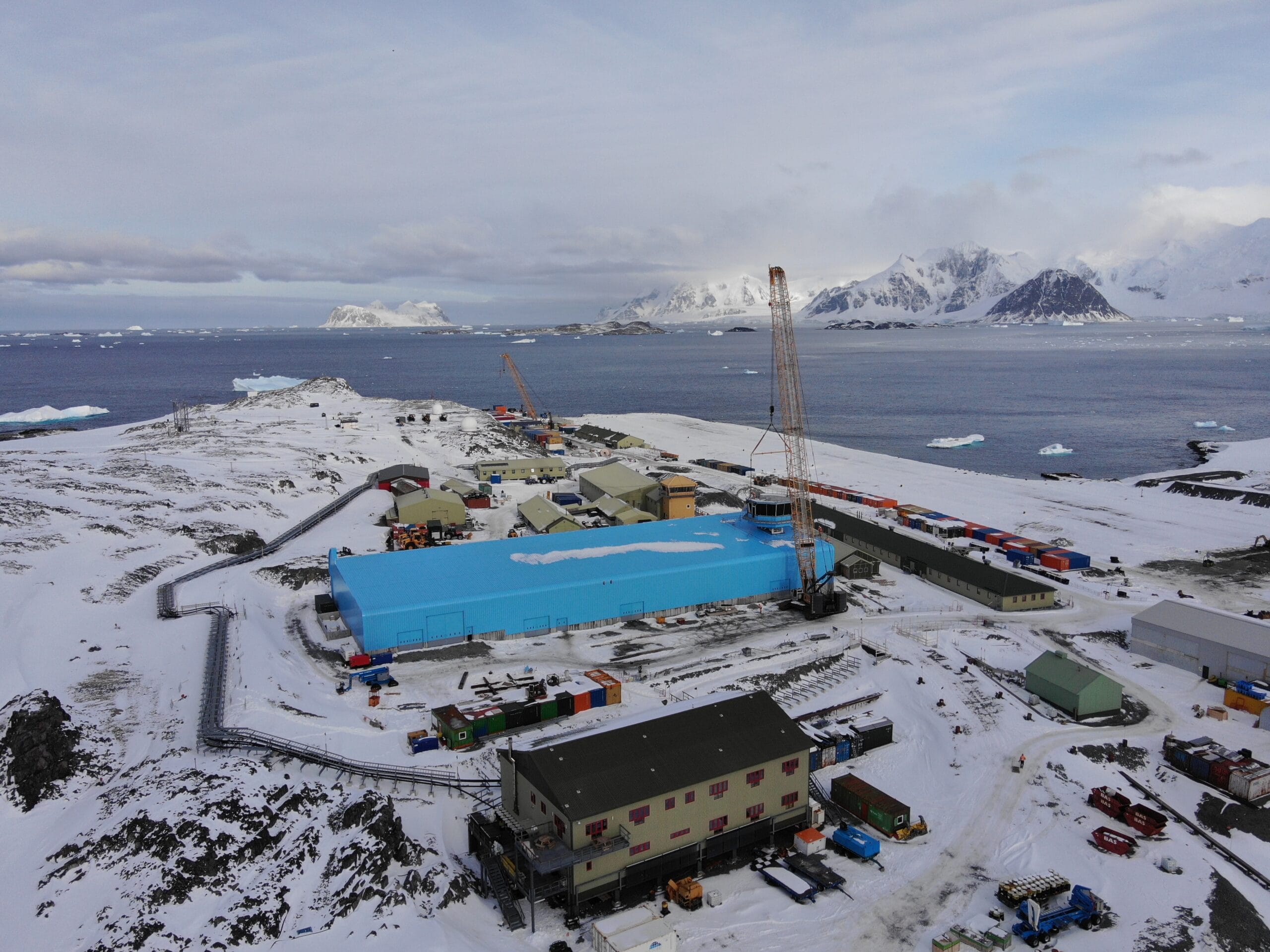 A view of a research village. There is a large blue building in the middle, with smaller green buildings surrounding it. The ground is covered with snow and there is open water and snowy mountains in the background.