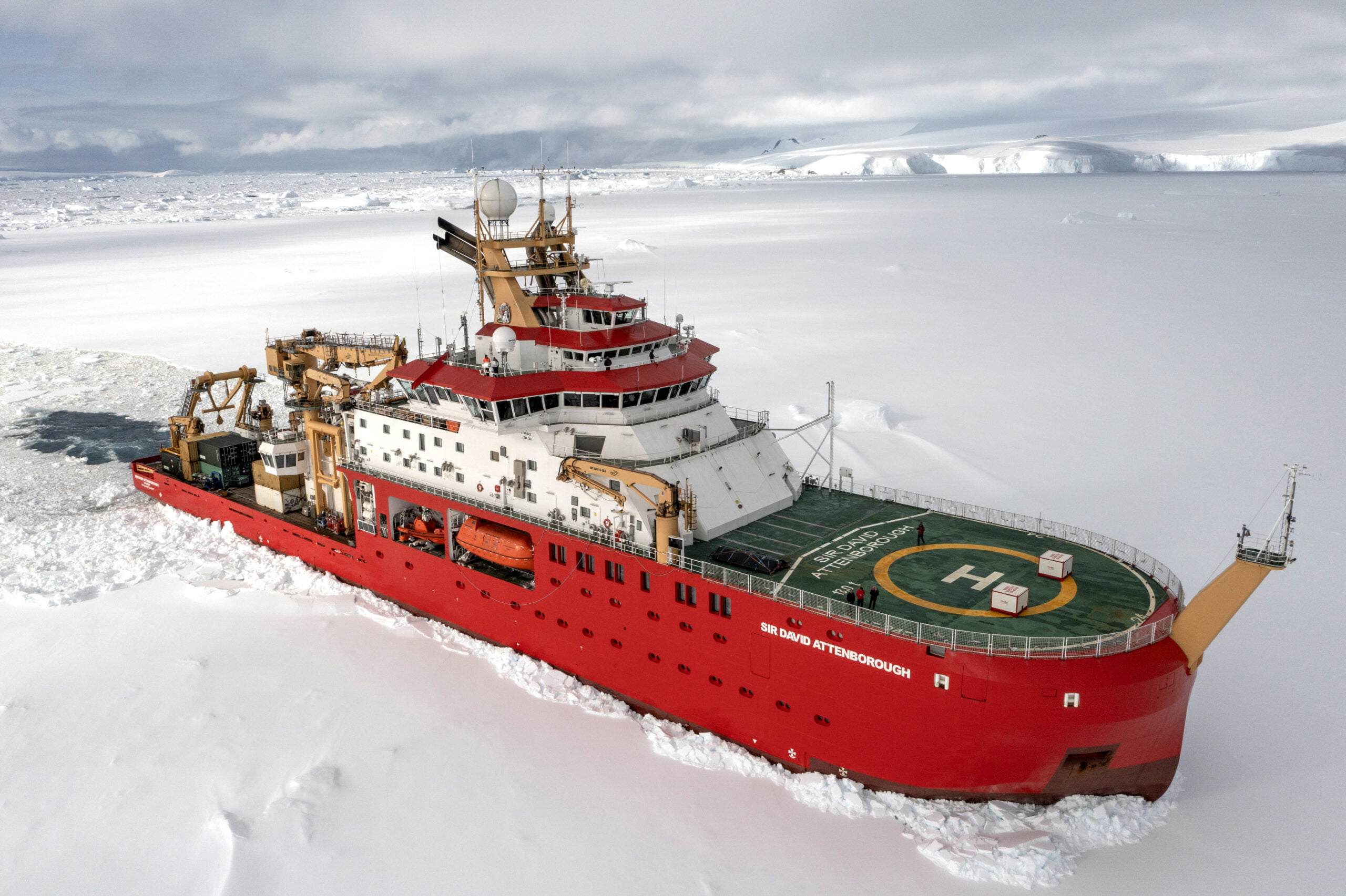 A large red ship breaking through ice. The ship has a large green helideck at the front, and the upper decks are white. There are yellow cranes and winches on the aft deck. The ship has the name Sir David Attenborough written in large white letters along the bow.