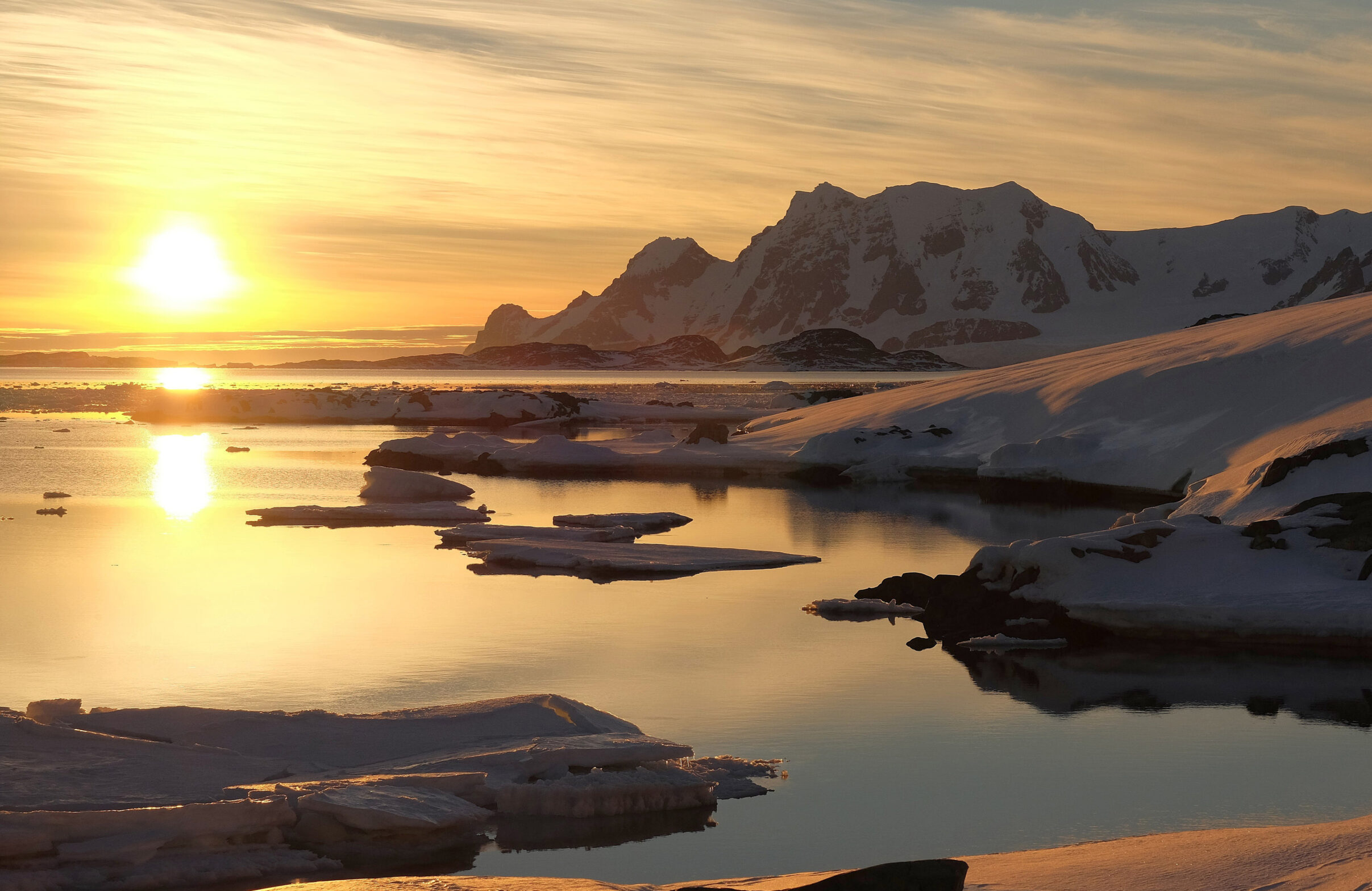 A photo of the sun setting over the ocean with snow covered mountains on the right