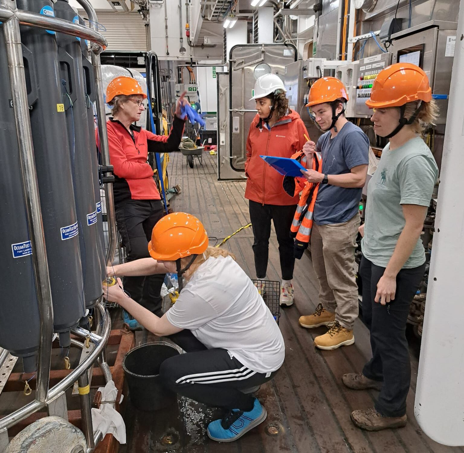 A group of people wearing hard hats on the deck of a ship, watching a woman talk. A person crouches down collecting water samples from a piece of science kit.