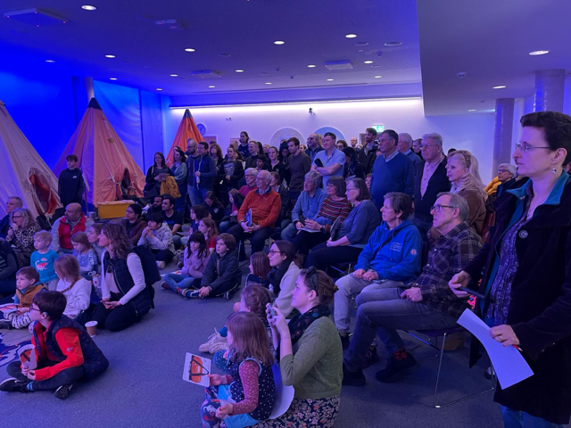 A large group of people sat in a room looking forwards at someone speaking.