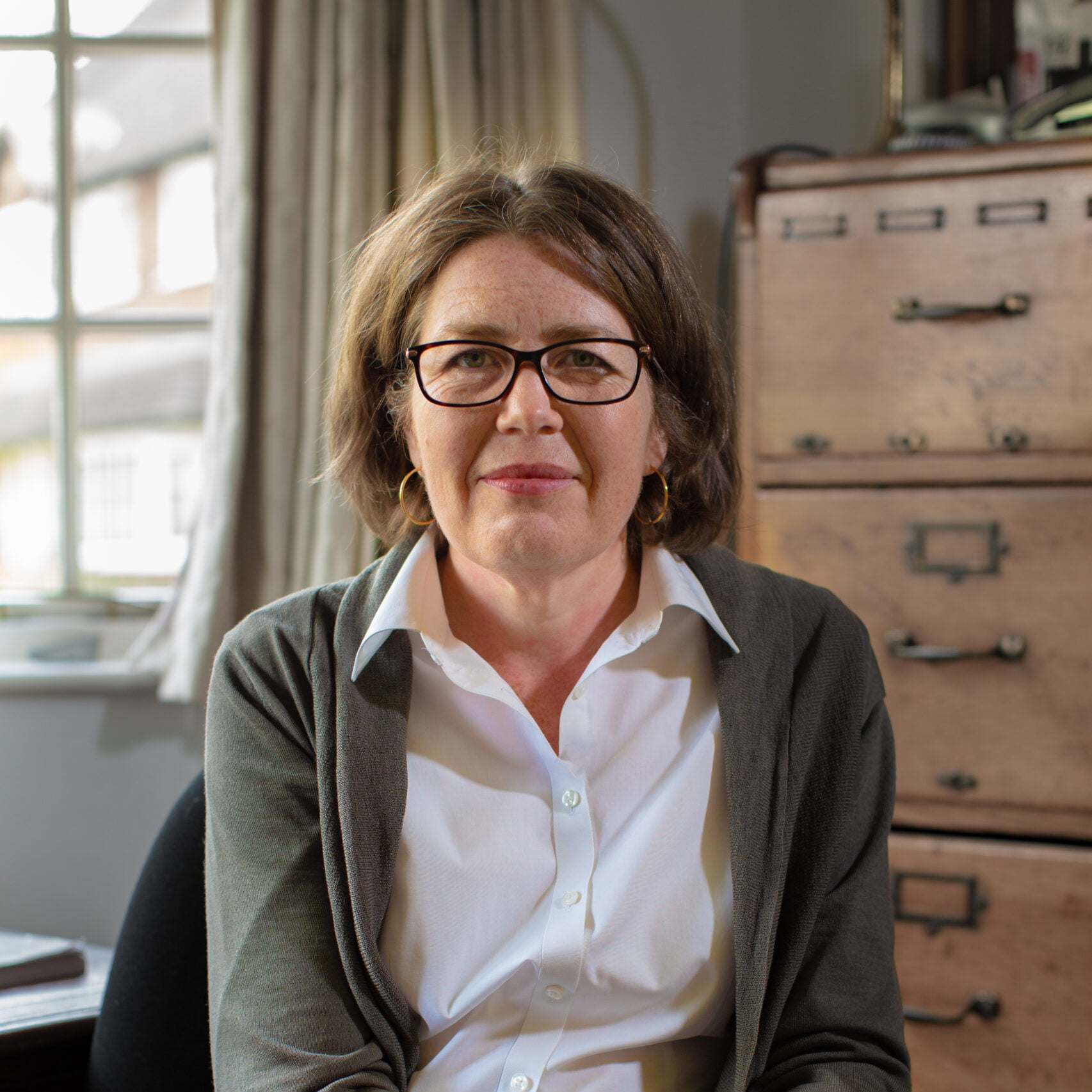 A woman with glasses sat on a chair with a window and large wooden chest of drawers in the background, smiling.