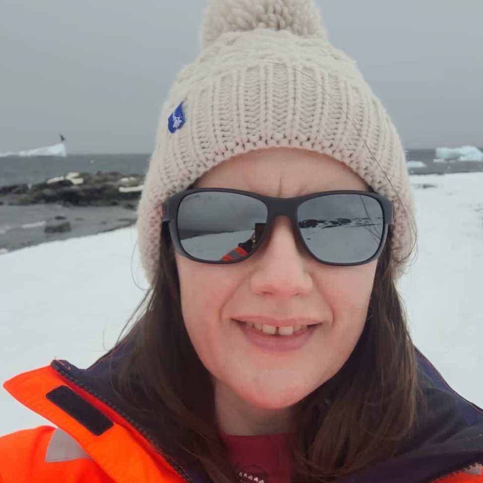 Elen is standing in Antarctica with Rothera Bay in the background. She is a white woman with brown hair, wearing a cream bobble hat and sunglasses.