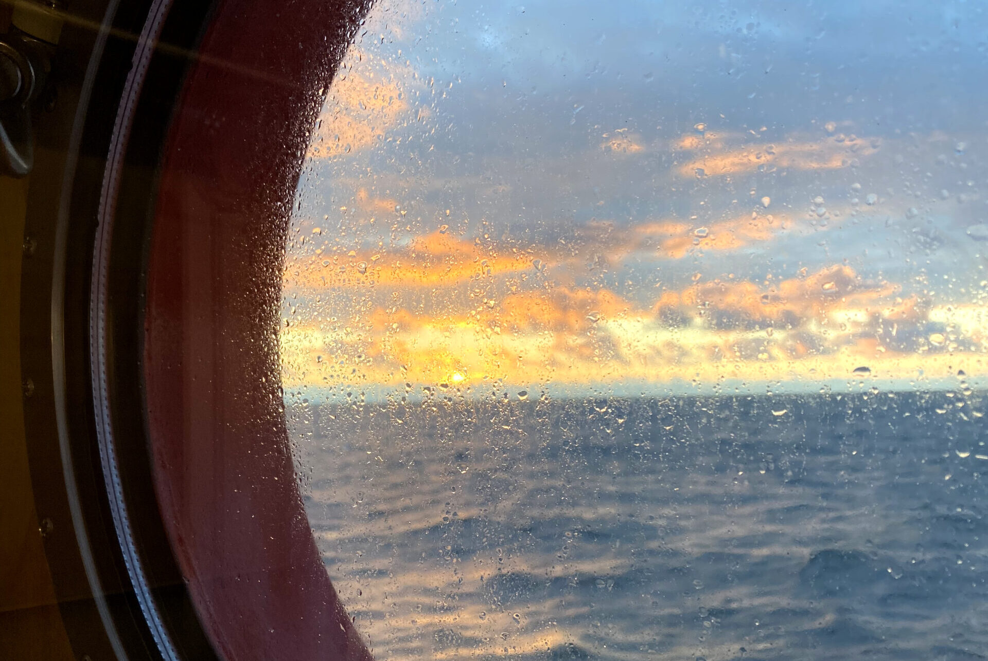 A view of the sunsetting over the ocean through a ship’s porthole. There is sea spray on the glass so the sunset and ocean are out of focus