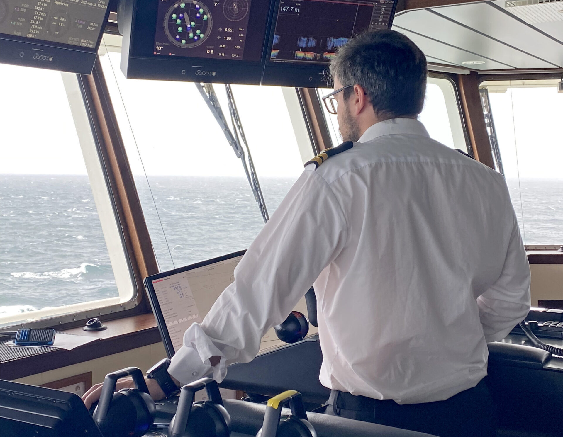 A man wearing a shirt, with his back to the camera, is looking at navigation equipment on the Bridge of a ship