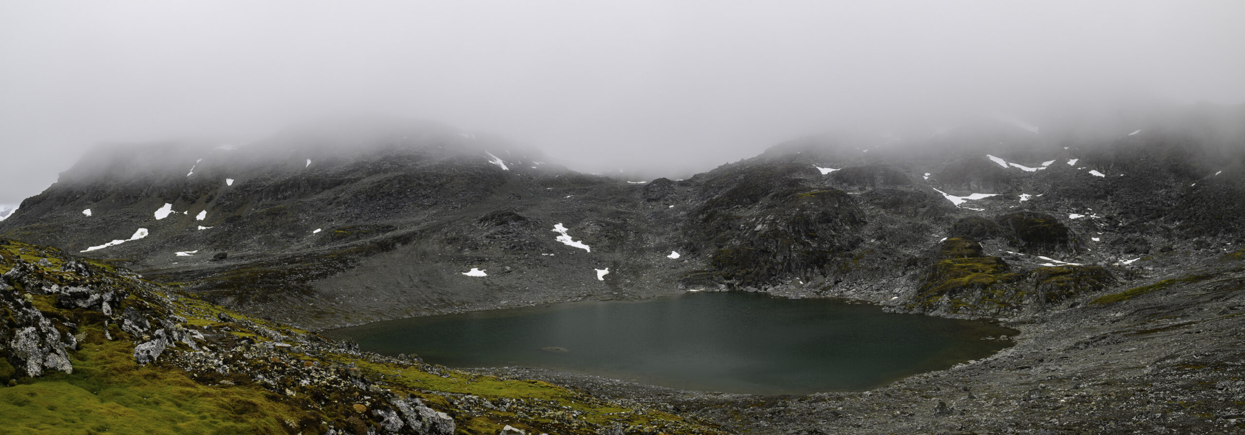 A panoramic photo of a deep green lake surrounded by low rocky mountains and low cloud