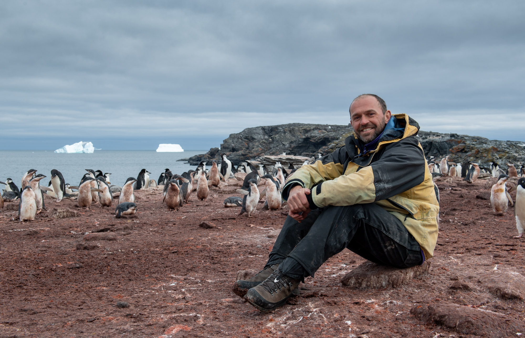 A smiling man sat on the ground, with penguins behind him