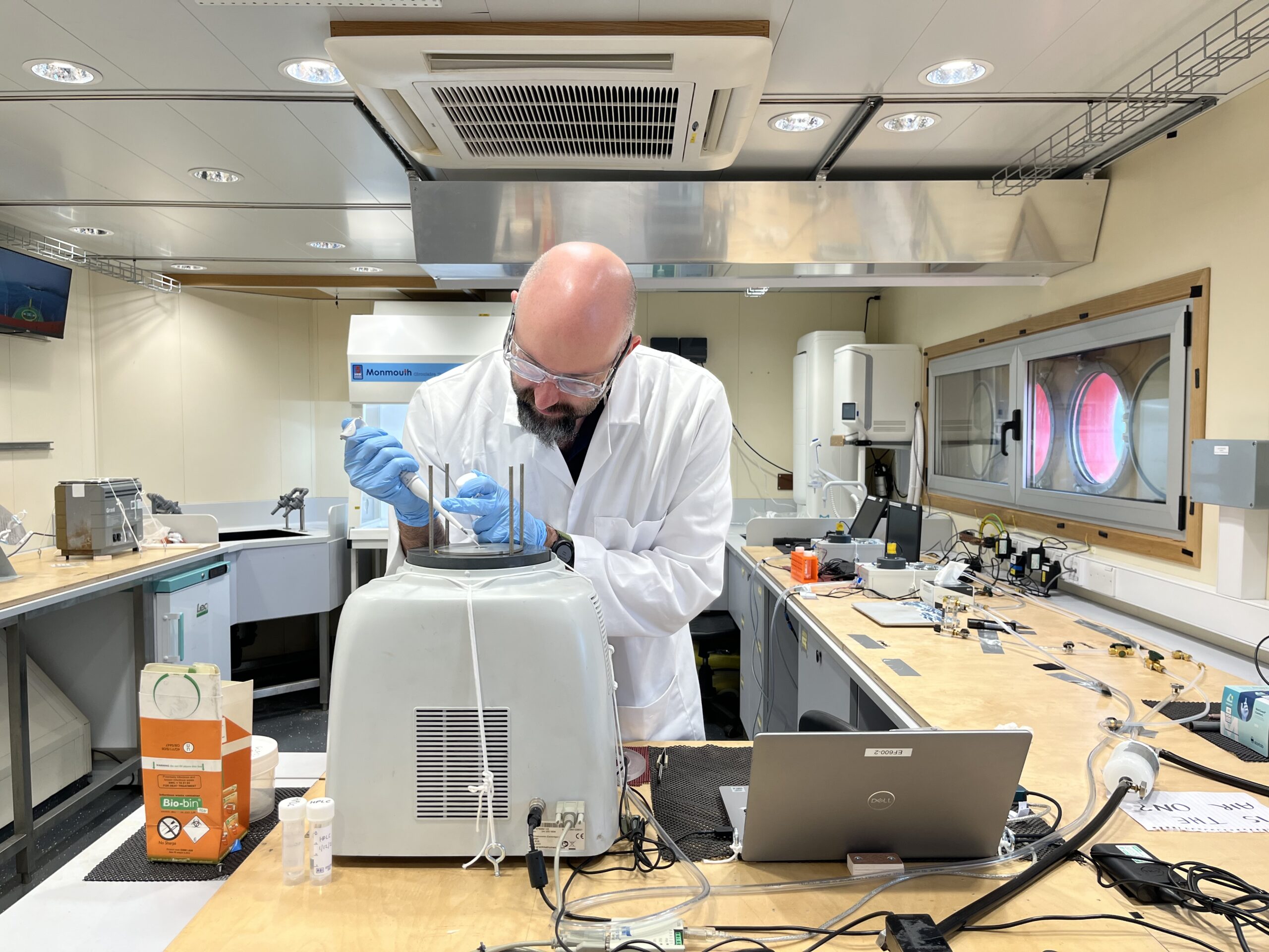 A man with a beard in a white lab coat and blue gloves. There is a lap top and scientific instrument in front of him. He is pipetting a sample.