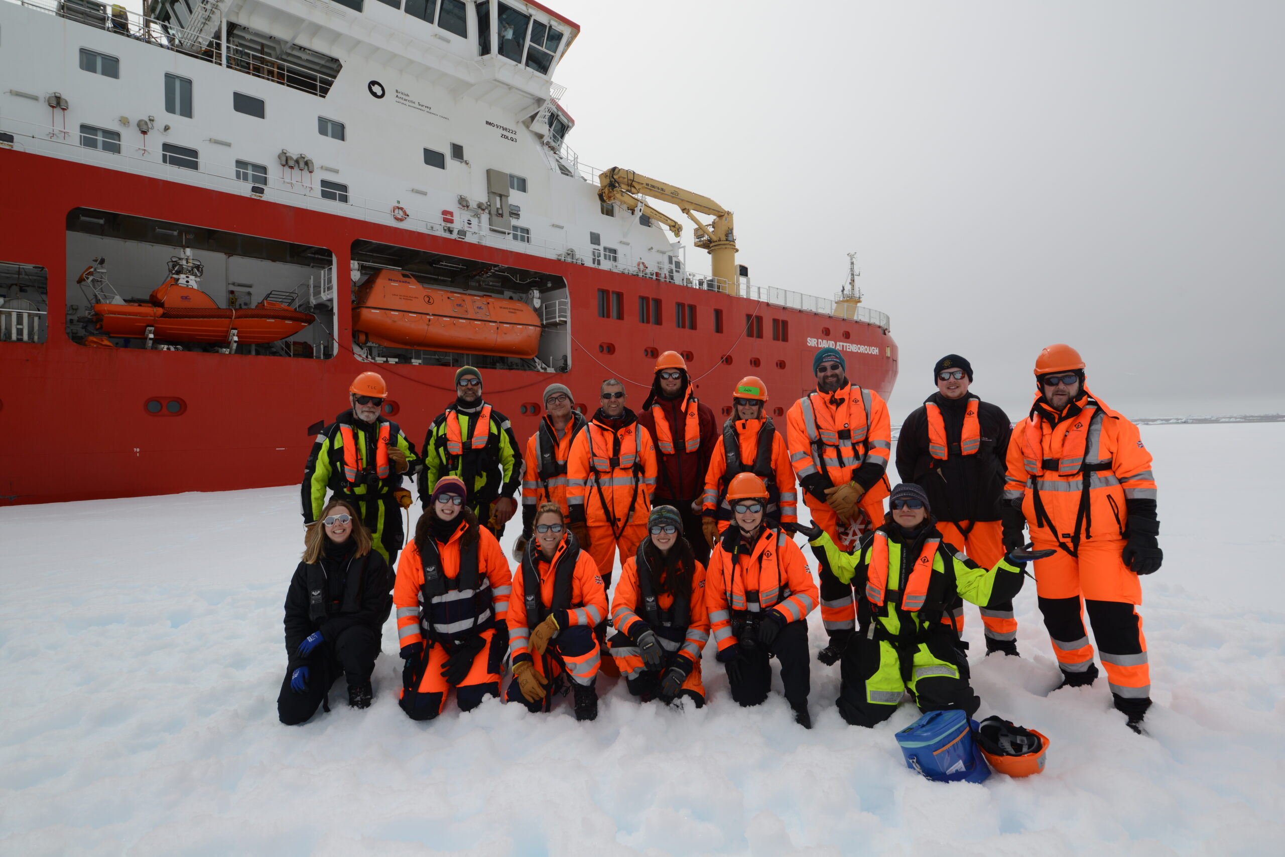 A group of people in high-vis outdoor gear, stood on sea ice with a red ship behind them