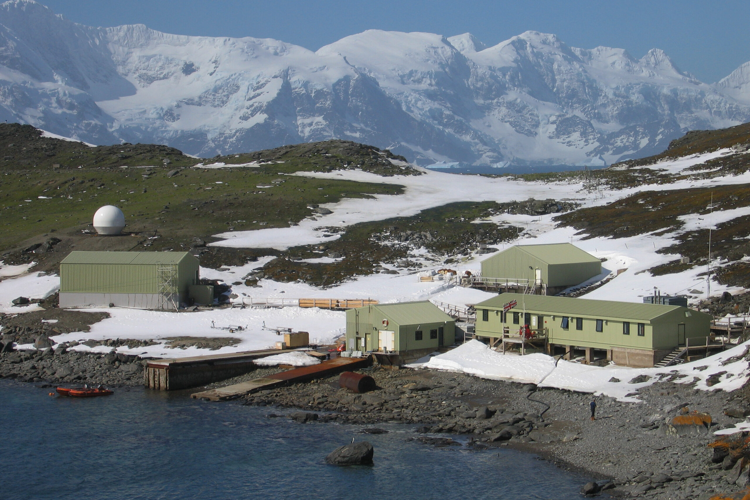 A collection of light green buildings on the water's edge, with snow covered mountains behind