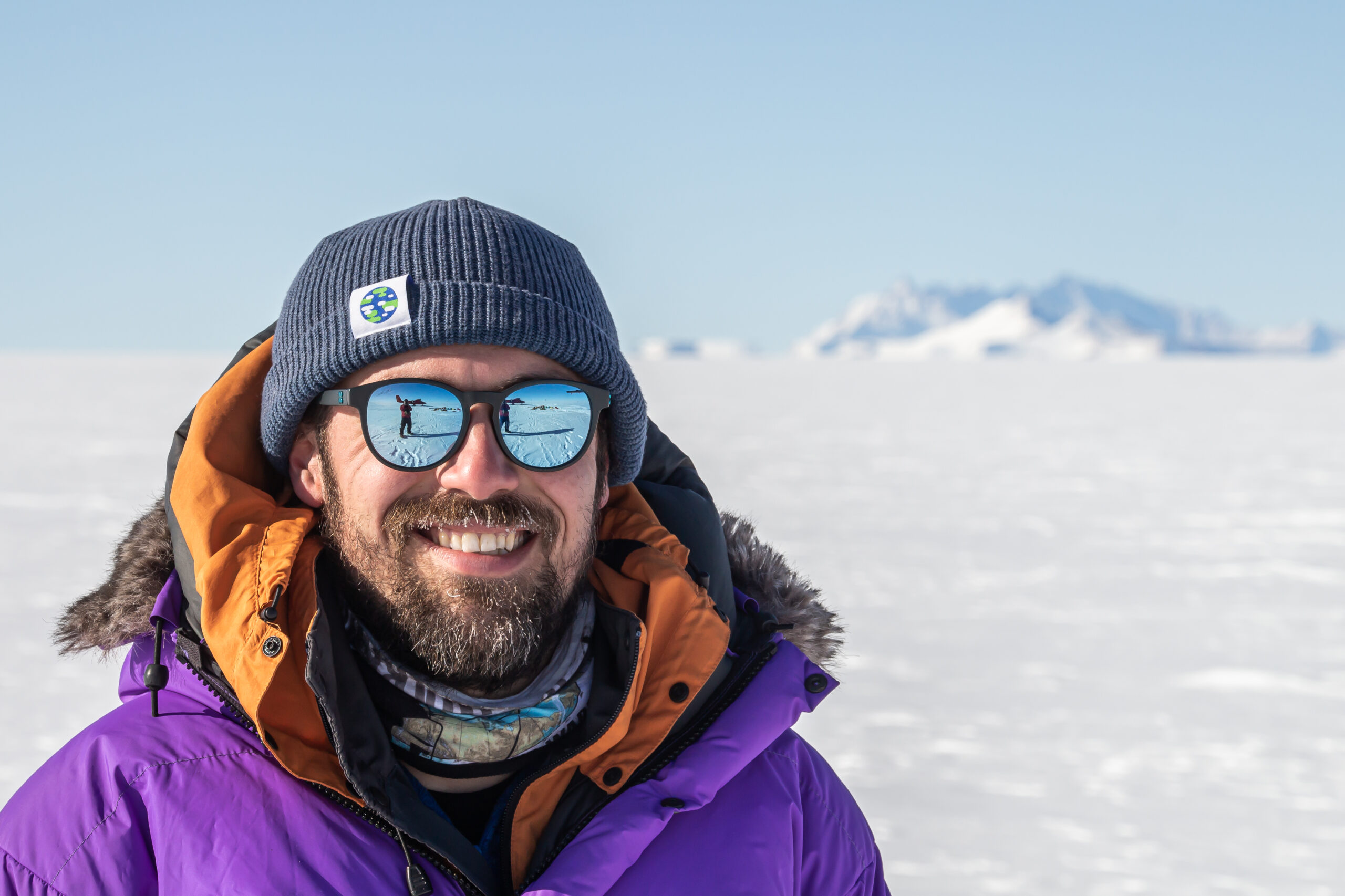 A man in lots of thick coats, hat and sunglasses in a sunny, snowy scene