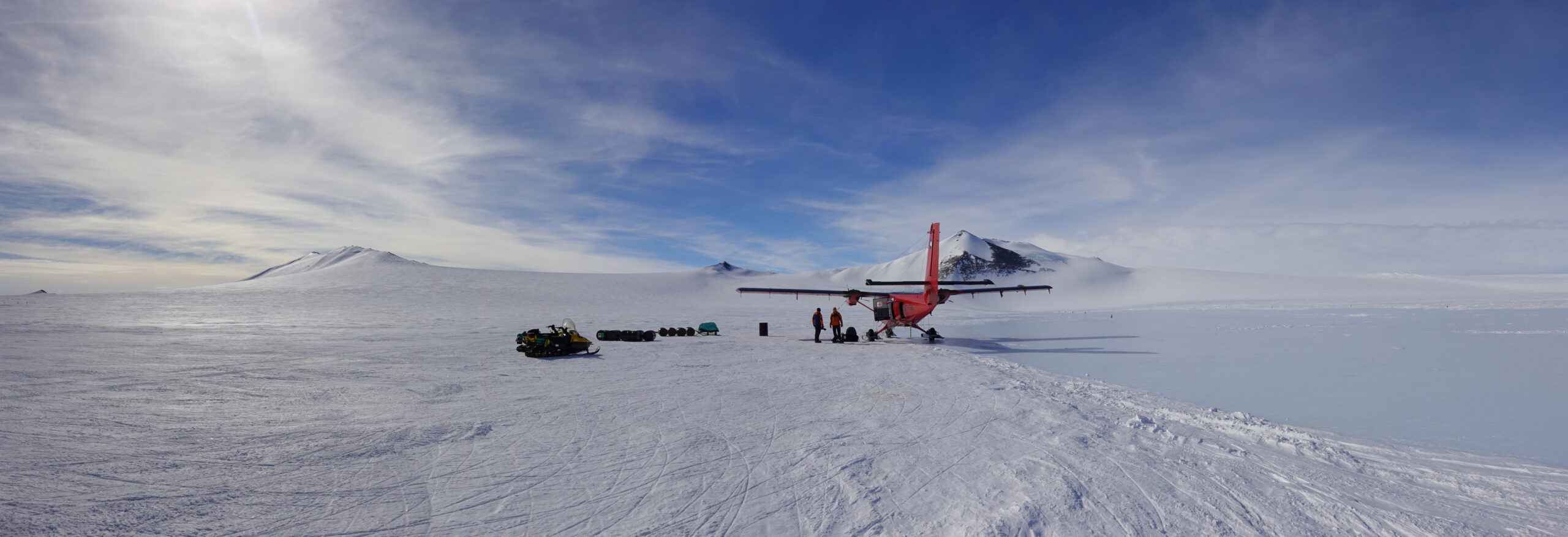 Two people stood under the wing of a small red plane in a snowy landscape. There are blue skies and a snow covered mountain in the distance