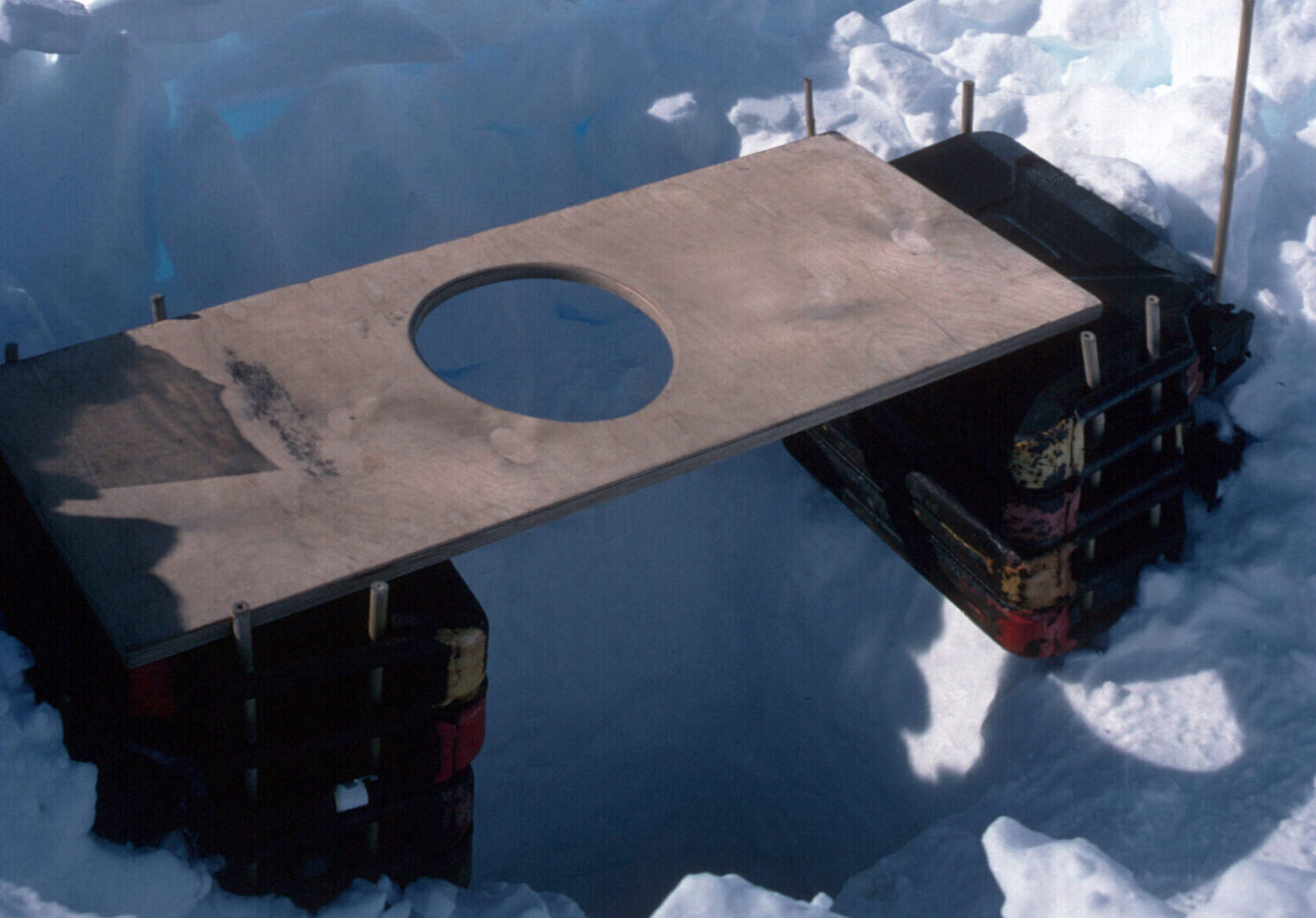 A wooden plank with a hole in the middle, balanced on a stack of boxes, over a deep hole dug in the ice