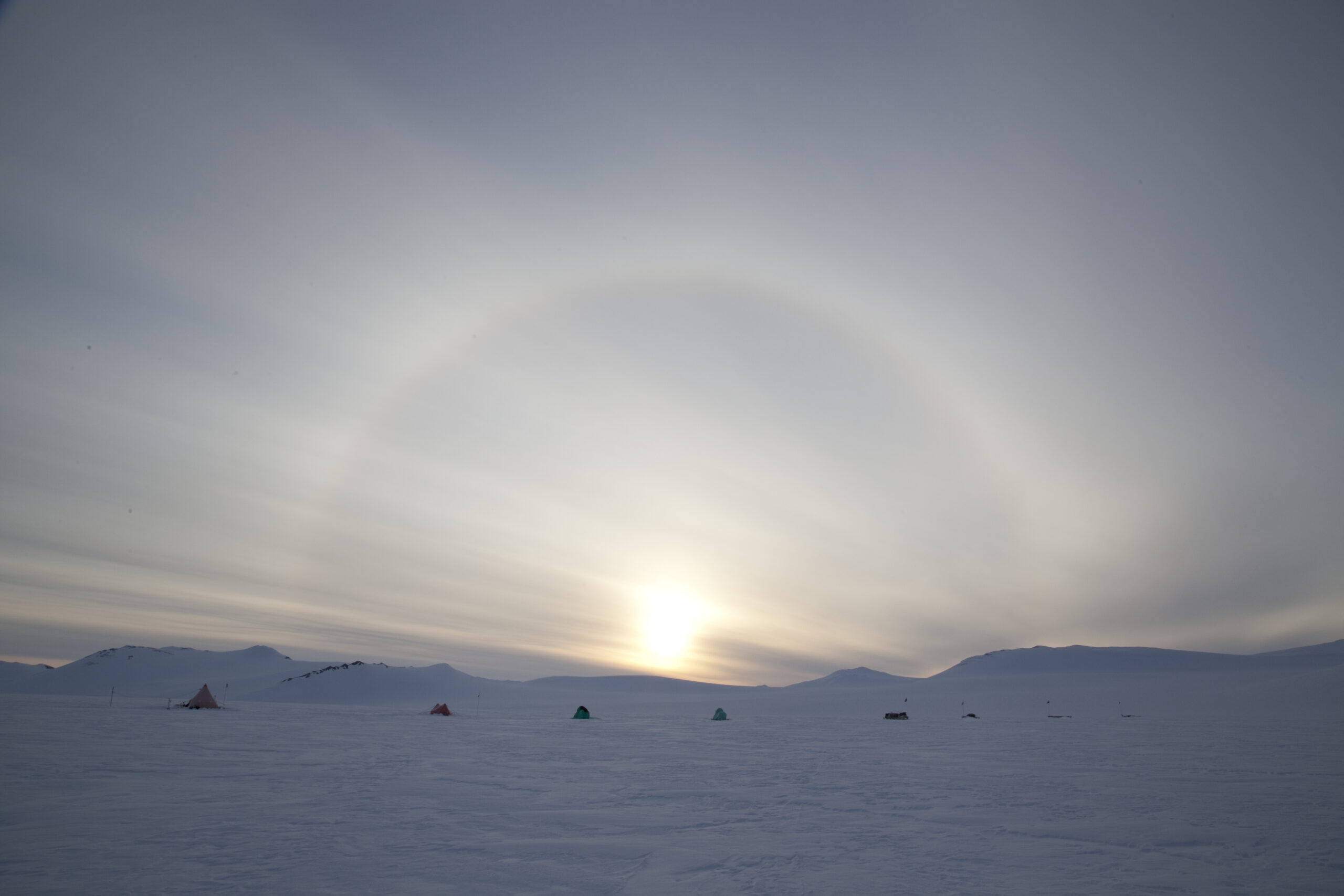 A photo of a light phenomenon called a sun halo - a halo of light arches over a sun sitting low in the sky, above a vast snowy landscape
