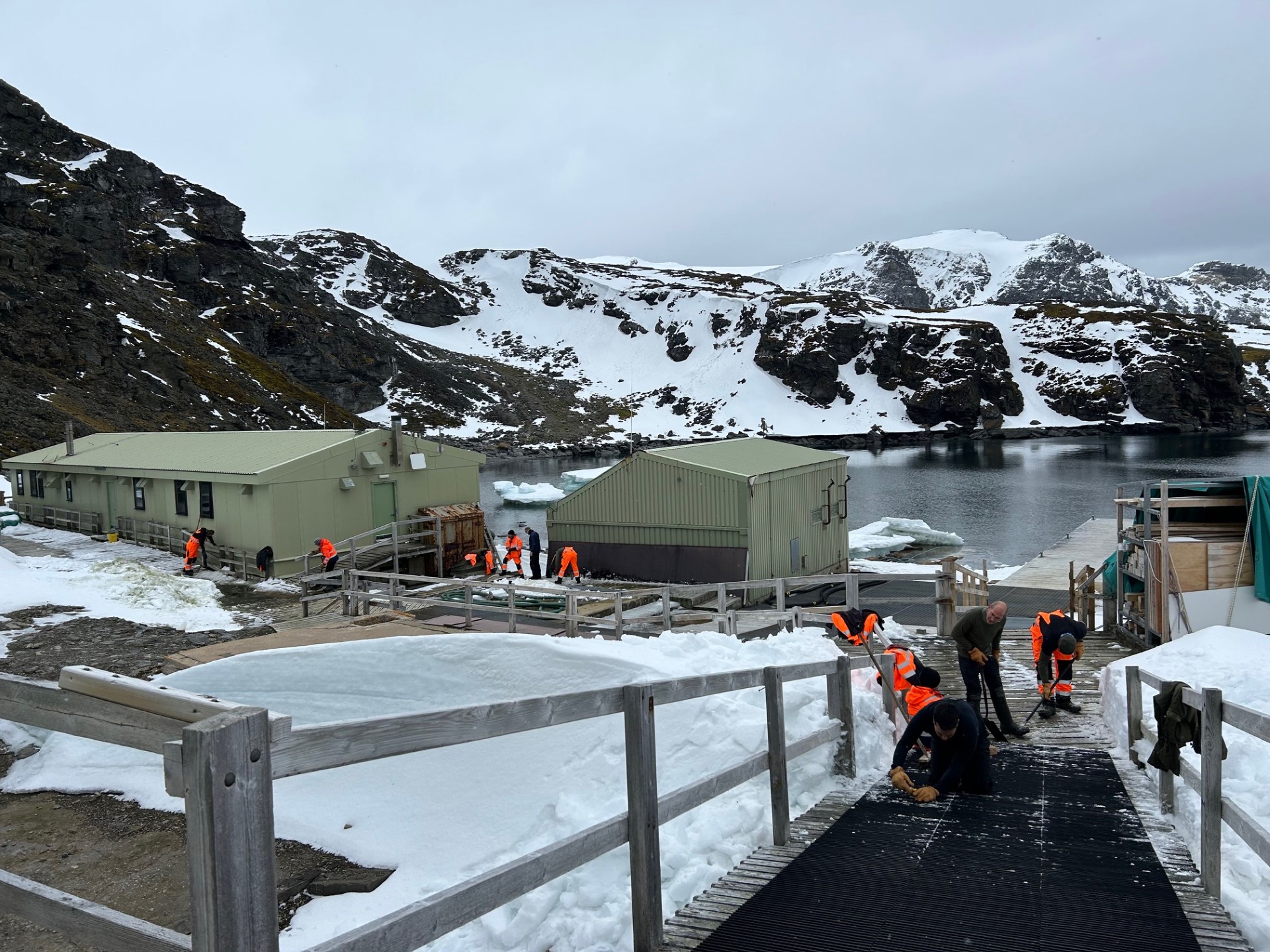 A group of people in high-vis jackets clearing ice from a sloped walkway down to some metal green huts. There are snow covered mountains in the background