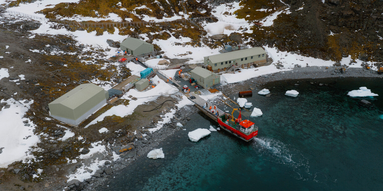 An aerial shot of a small red workboat berthed up near four green metal buildings in a snowy landscape