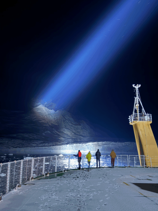 People stood on the helideck of a ship in the dark, looking at a glacier illuminated by the ship's searchlight