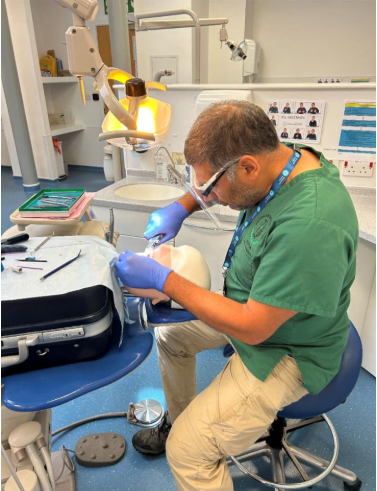 A man in medical clothes sits on a stool carrying out a dental procedure on a dummy