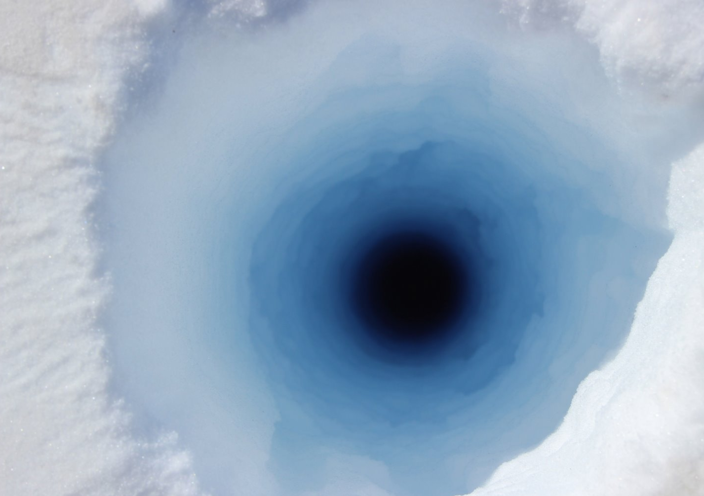 Looking down a narrow but deep hole drilled into the ice sheet. The colour changes from white to shades of blue and then black