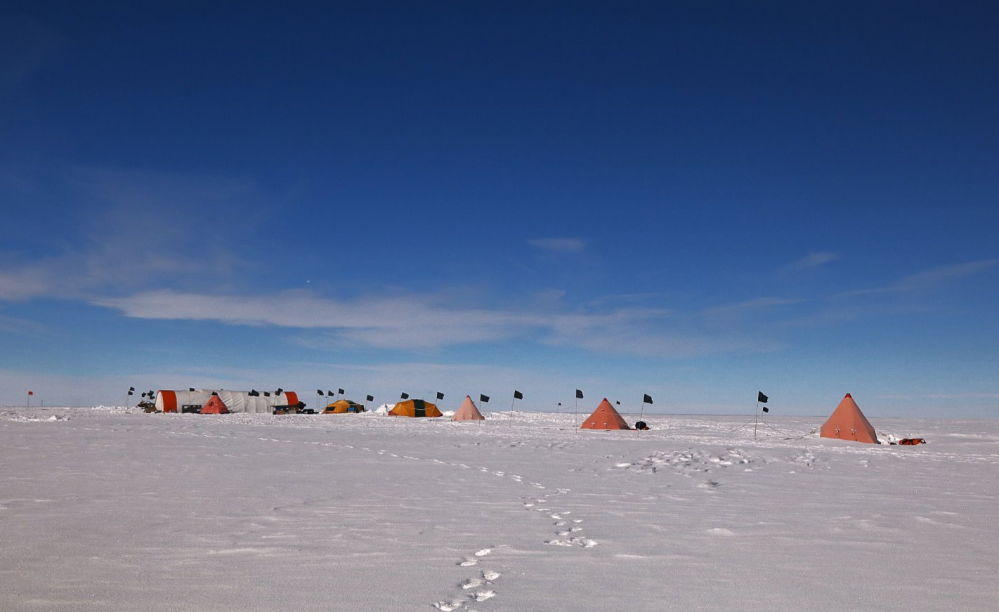 A group of tents in a line, in a snowy landscape with blue skies. There are also lots of flag markers.