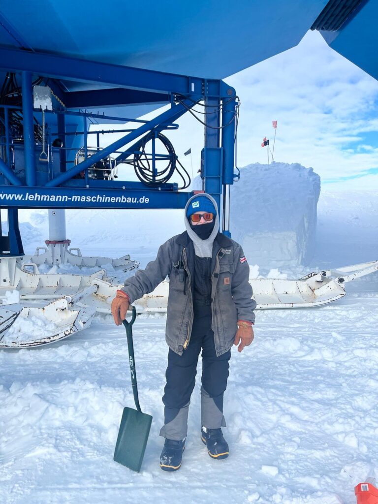Person dressed in winter gear stands on snowy terrain, holding a shovel. They are near a large blue machine, with ice equipment in the background. The scene feels cold and industrious.