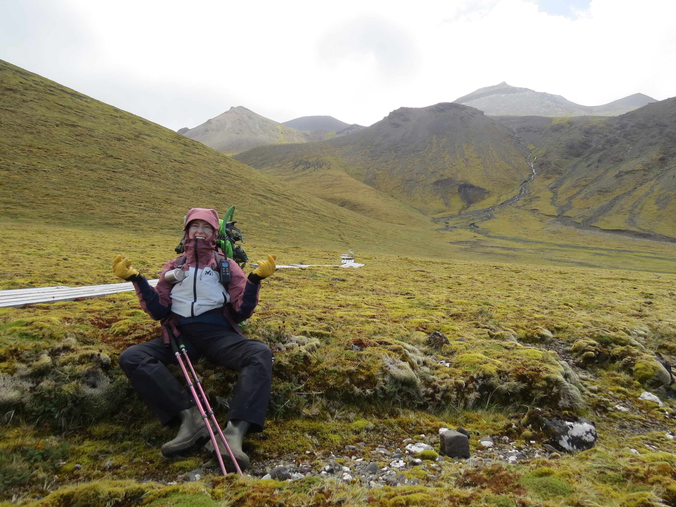 A hiker with trekking poles sits smiling on a grassy hillside, wearing a pink jacket, yellow gloves, and a backpack. Behind are rolling green hills and a cloudy sky.