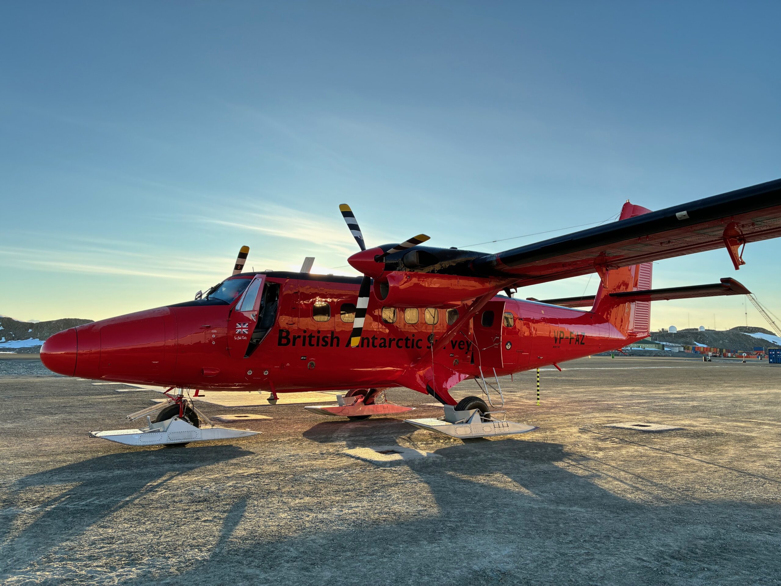 Red British Antarctic Survey plane on a sunny tarmac in Antarctica. The sun casts long shadows, with mountains and blue sky in the background.