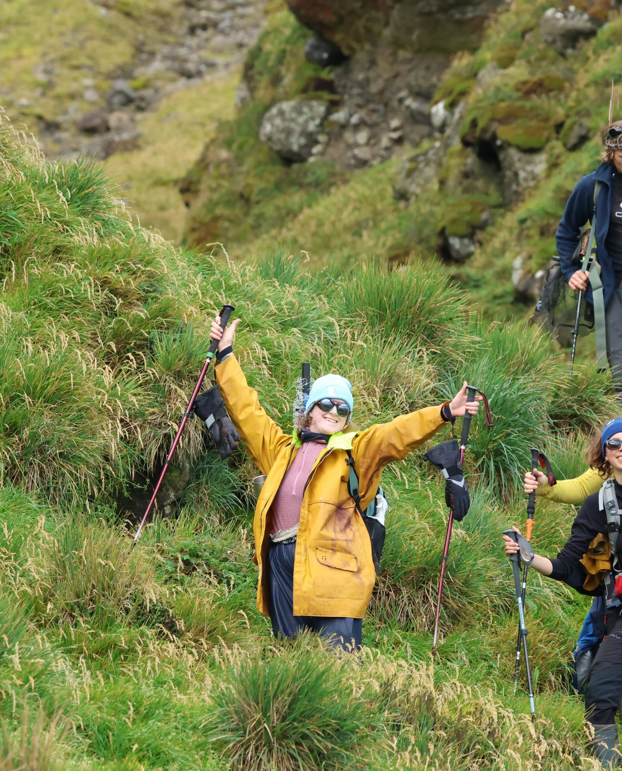 A smiling hiker in a yellow jacket and sunglasses stands with arms outstretched on a green, grassy hillside, radiating joy and accomplishment.