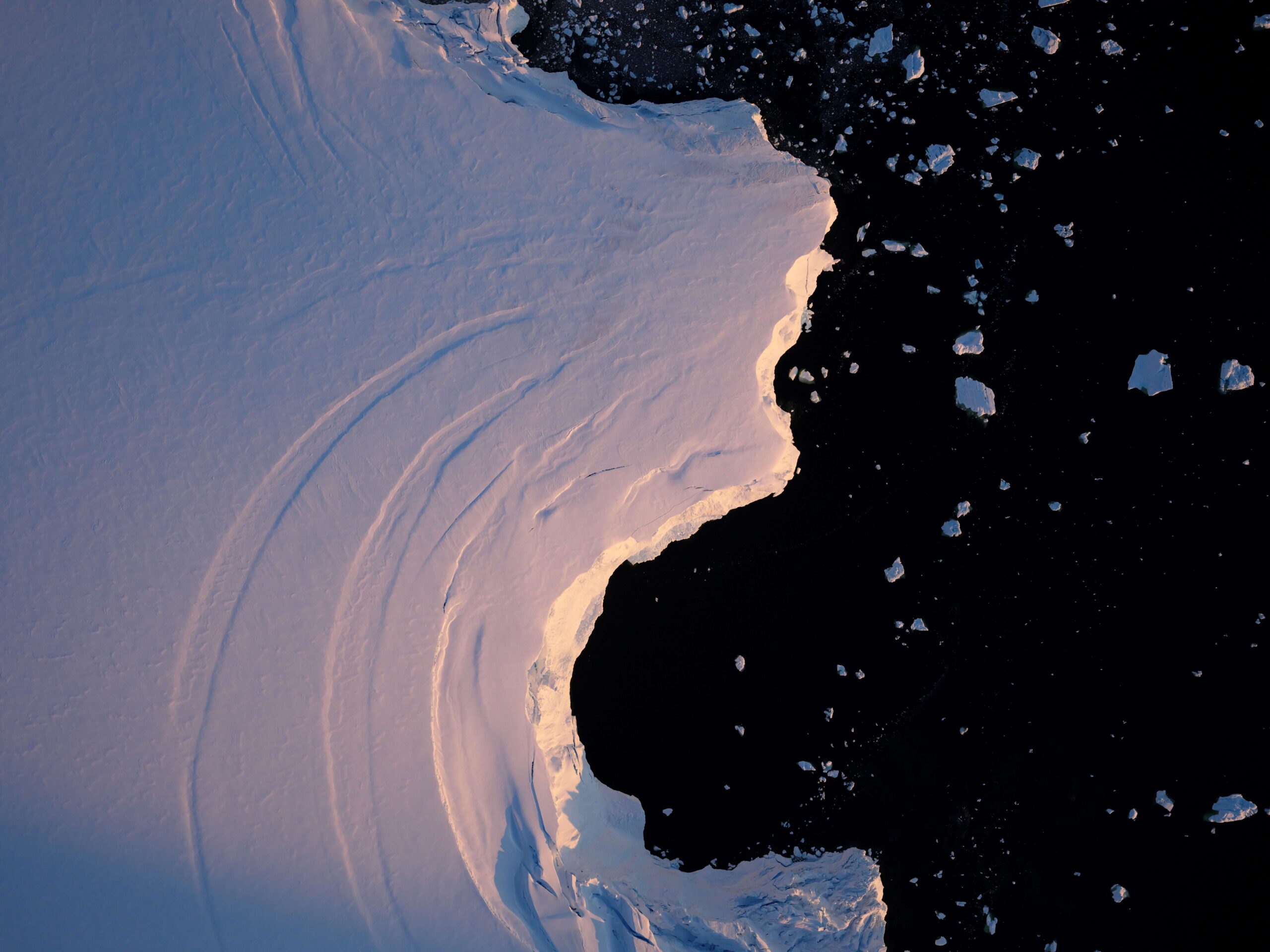 Aerial view of snow-covered land partially illuminated by sunlight, contrasting sharply against dark, icy water with scattered ice chunks. Serene and dramatic.